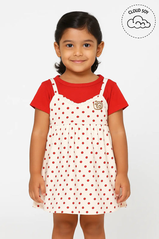 A young girl with dark hair pulled back smiles, wearing a short-sleeved red top underneath a sleeveless, cream-colored dress with small red polka dots. The dress has thin straps and a small embroidered patch on the chest. A "Cloud Soft" logo is visible in the upper right corner. This is a cute piece of girls kids clothing and trending clothes for girls, showcasing a sweet style in girls fashion. It's a lovely dress for kids girls.