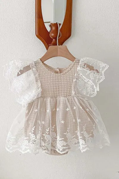 A delicate, cream-colored lace romper with flutter sleeves, hanging from a wooden hanger against a white wall with a partial view of a wooden mirror frame.