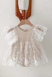 A delicate, cream-colored lace romper with flutter sleeves, hanging from a wooden hanger against a white wall with a partial view of a wooden mirror frame.