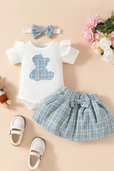 A stylish set of new born cloths for a baby girl, including a white ribbed bodysuit with a blue teddy bear, a blue and white plaid skirt, and a matching headband, showcasing trending clothes for girls fashion.
