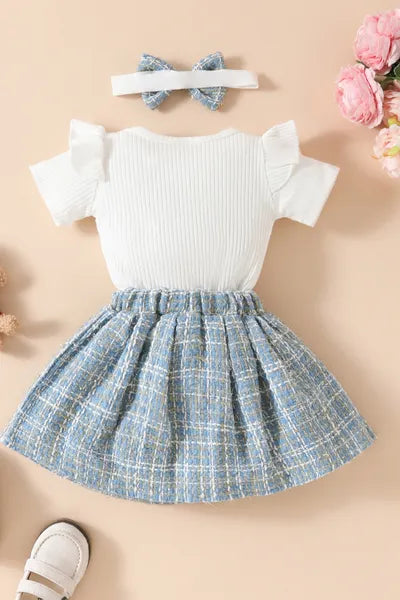 A sweet ensemble of new born cloths for a baby girl: a white bodysuit with a teddy bear, a blue plaid skirt, and a headband, reflecting current trends in girls fashion. While the top is a bodysuit, the overall look could be considered a type of romper dress.
