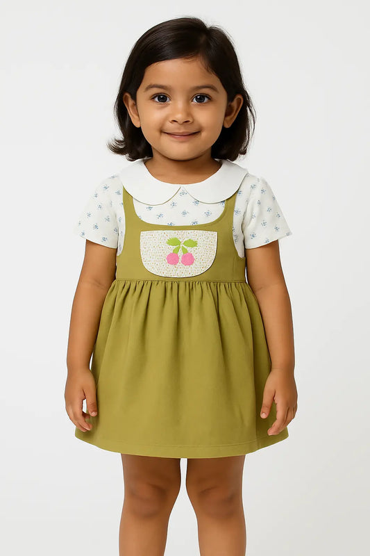 A young girl with short dark hair smiles, wearing a short-sleeved white top with a subtle floral print and a rounded collar underneath an olive green overall dress. The dress has a cream-colored bib pocket with embroidered pink cherries. This is a charming piece of girls kids clothing and trending clothes for girls, showcasing a sweet style in girls fashion. It's a lovely dress for kids girls.