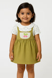 A young girl with short dark hair smiles, wearing a short-sleeved white top with a subtle floral print and a rounded collar underneath an olive green overall dress. The dress has a cream-colored bib pocket with embroidered pink cherries. This is a charming piece of girls kids clothing and trending clothes for girls, showcasing a sweet style in girls fashion. It's a lovely dress for kids girls.