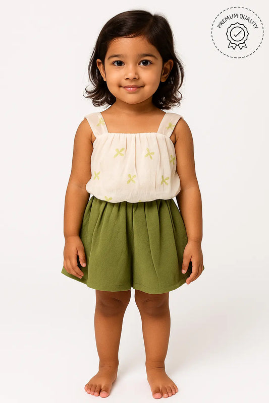 A young girl with short dark hair smiles, wearing a sleeveless cream-colored top with small light green embroidered details and a pair of olive green shorts. A "Premium Quality" badge is visible in the upper right corner. This is a cute outfit in girls kids clothing and trending clothes for girls, showcasing a stylish look in girls fashion. It's a lovely set of clothes for kids girls.