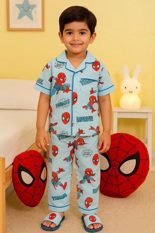 A young boy smiles while wearing a light blue pants set - boys pyjamas featuring a Spider-Man print with red piping. He is standing in a bedroom setting with Spider-Man-themed pillows and blue slippers, showcasing kids sleepwear from the boys nightwear collection.