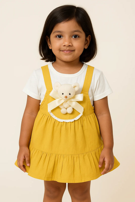 A young girl models a cute mustard yellow overall dress with a white t-shirt underneath, showcasing a sweet look in girls fashion. The dress features a small white teddy bear with a striped ribbon, making it a charming piece of girls kids clothing and trending clothes for girls. This is a lovely example of clothes for kids girls.