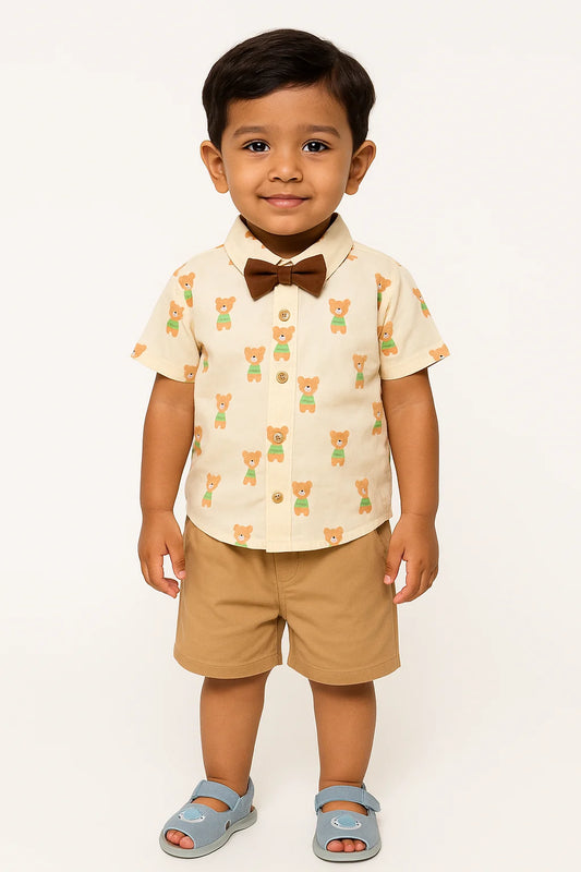 A young boy with dark hair smiles while wearing a short-sleeved, collared shirt with a teddy bear print, paired with khaki shorts, a brown bow tie, and light blue sandals. He is standing against a white background.