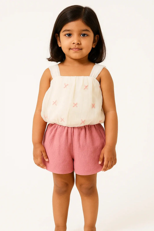 A young girl with short dark hair smiles at the camera, wearing a sleeveless cream-colored top with small pink embroidered details and a pair of soft pink shorts. She is standing against a plain white background.