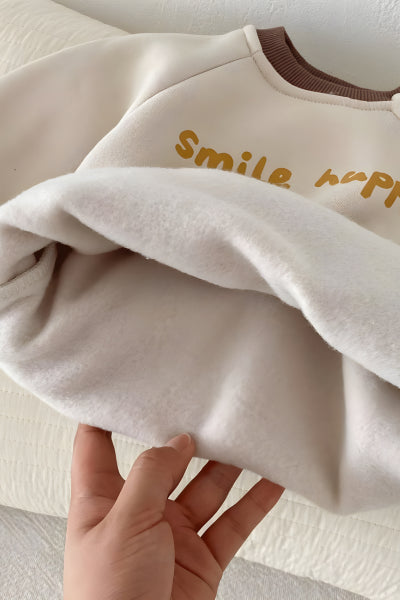 Beige baby sweater with 'Smile happy' text held by a hand on a light background