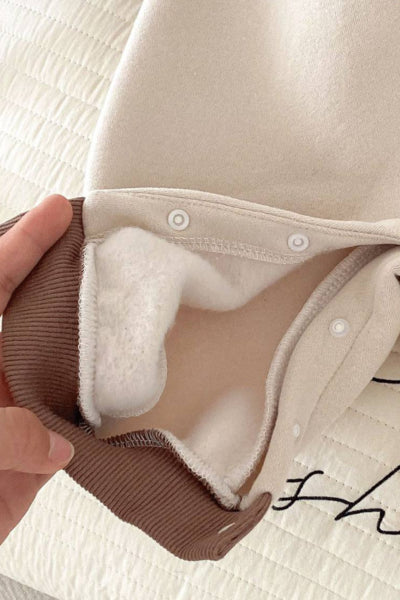 Close-up of a beige garment with brown trim held by a hand on a light background.