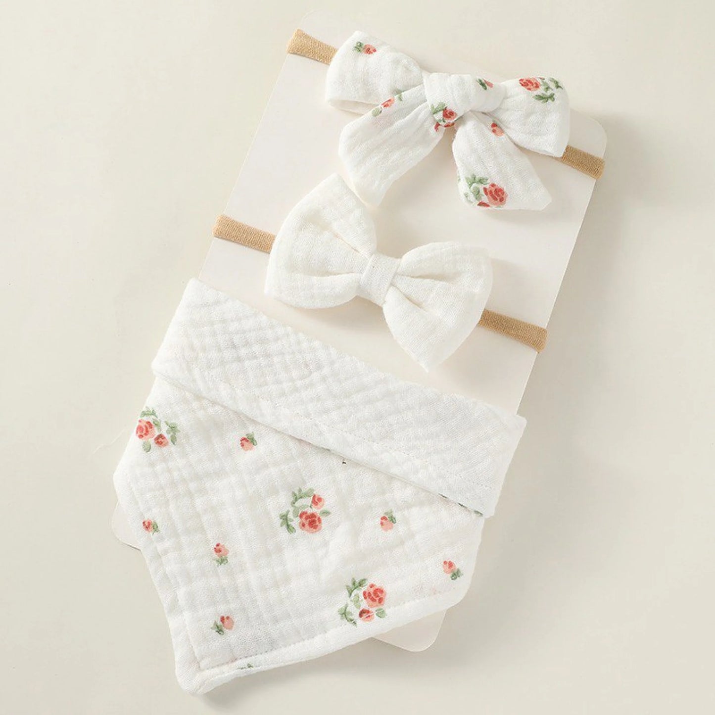 A set of three white baby accessories: two headbands with small white bows (one with a subtle floral print) and a bandana bib with a matching small floral print, arranged on a light beige surface.
