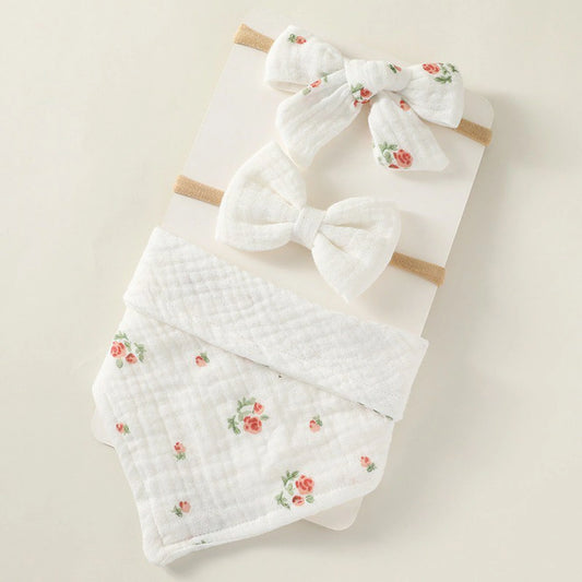 A set of three white baby accessories: two headbands with small white bows (one with a subtle floral print) and a bandana bib with a matching small floral print, arranged on a light beige surface.