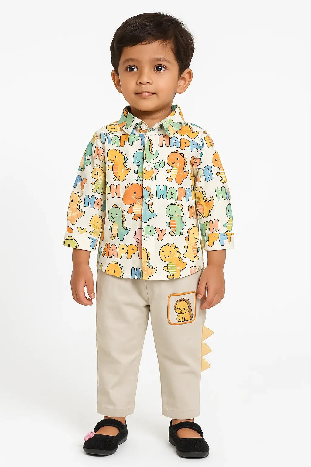 A young boy with dark hair stands against a white background, wearing a long-sleeved button-down shirt with a colorful dinosaur print, light beige pants with a dinosaur patch and tail detail, and black Mary Jane shoes.