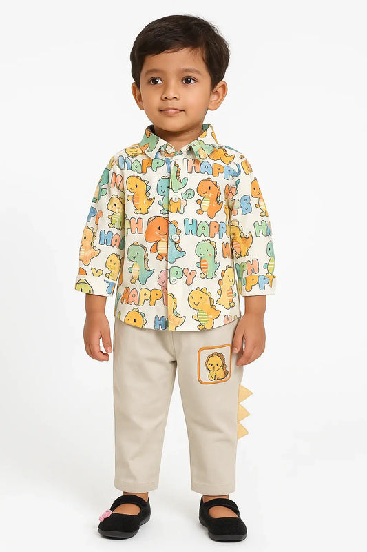 A young boy with dark hair stands against a white background, wearing a long-sleeved button-down shirt with a colorful dinosaur print, light beige pants with a dinosaur patch and tail detail, and black Mary Jane shoes.