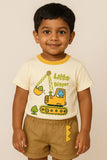 A young boy with dark hair smiles while wearing a cream-colored short-sleeved t-shirt with a yellow trim and a cartoon excavator graphic that reads "Little Digger." He is also wearing khaki shorts and standing against a light beige background.